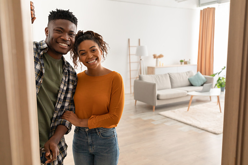 Joyful African American Family Couple Embracing Standing In Opened Doors Of Their New Home, Smiling Looking At Camera. Housing For Married Couple. Real Estate Property Ownership Concept
