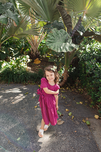 Cute Cuban-American 3-Year-Old Toddler Girl With Brown Curly Hair, Brown Eyes & Olive Skin Tone Wearing a Bright Purple Dress, a Hair Bow & White Sandals While in Front of Tropical Plants in Palm Beach, Florida