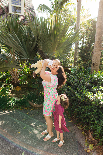 A Happy 31-Year-Old Cuban Mother with Curly Brown Hair & Wearing a Colorful Spring Dress While Holding Her Snuggly Baby Boy & Standing by Her 3-Year-Old Daughter While Walking Peacefully on a Lake Trail in Palm Beach, FL