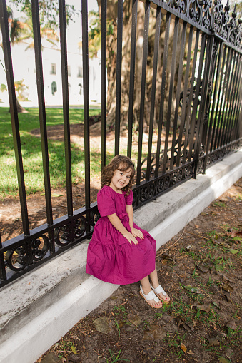 Cute Cuban-American 3-Year-Old Toddler Girl With Brown Curly Hair, Brown Eyes & Olive Skin Tone Wearing a Bright Purple Dress, a Hair Bow & White Sandals While in Front of a Black Railing on a Lake Trail in Palm Beach, Florida