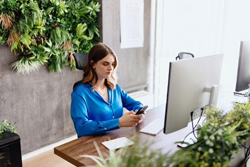 Young motivated businesswoman working from the modern office while using her black smartphone