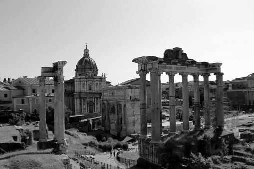 Rome, Italy – June 23, 2022: A grayscale shot of a historical scene featuring a clock tower atop a church surrounded by ancient ruins of the Roman Forum