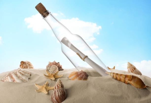 istock Corked glass bottle with rolled paper note and seashells on sand against sky 1468542186