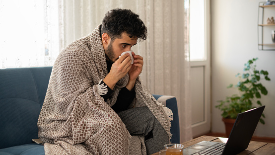 Man covered in warm blanket looking at laptop screen, working online from home, sneezing nose, middle-aged freelancer got sick, having cold, flu or coronavirus, copy space