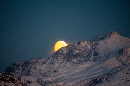 A near full moon is slowly creeping behind a mountain along the norwegian coast on an early winter morning near the town of Bodo.