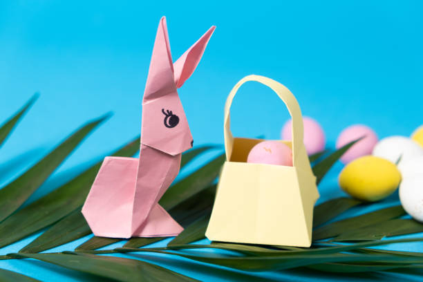 una statuetta del coniglietto pasquale fatta di carta, origami e un cesto con uova su sfondo blu, posto per il testo - figurine easter egg easter holiday foto e immagini stock