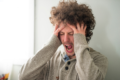 Millennial young man is holding his head tightly in anger and frustration, with his fingers through his long curly hair. He has an angry expression on his face as he is yelling out from stress. He is dealing with mental health issues and stress from economic pressure.