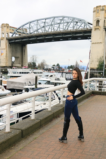 A Colombian model posing under the Burrard Bridge in Vancouver, BC. She has log, brown, straight hair with a long sleeve black top, jeans and knee high boots.