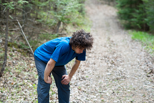 Man with curly brown hair in athletic clothing is bent over with his hands on his knees as he breathes heavily from exhaustion after running through a forest trail to exercise and improve his mental health.