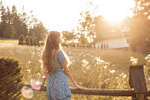 Rearview of carefree ginger woman in blue summer dress stand near wooden fence near house in sunny lawn in countryside.