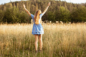 Red haired relaxed woman in blue summer dress stand with raised arms in sunny field with flowers near woodland. Freedom