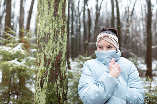 Portrait of mysterious middle-aged woman wearing beige hair band, covering face with blue warm puffy jacket, standing outside among trees in park forest in snowy winter. Winter activities, snowfall.