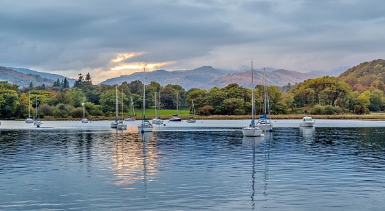 Sunset over Lake Windermere, Cumbria, UK with the mountains of the Lake District behind.  Yachts are moored at the edge of the lake and the area is popular with sailors, walkers and holidaymakers.