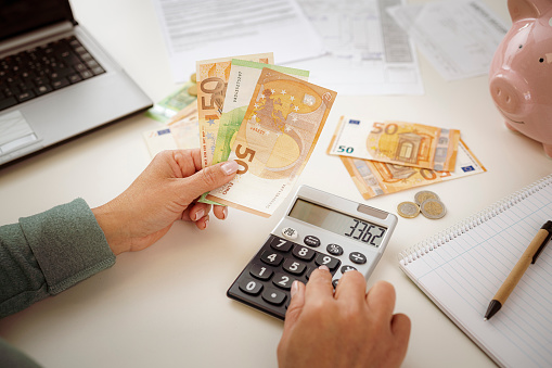 Close-up of woman's hands using calculator for planning home finances. She is holding Euro banknotes.