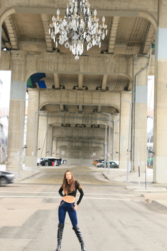 A Colombian woman posing under a huge chandelier hanging on a bridge in Vancouver BC. She is wearing long brown straight hair, a black long sleeved short top, jeans and knee high boots.