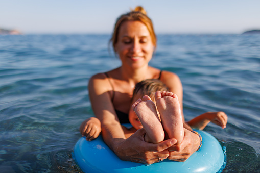 A cute young mother with blond hair smiles and shows to the camera the small tender legs and arms of her baby son, who is sitting in a bright blue inflatable ring in the blue dark Adriatic Sea