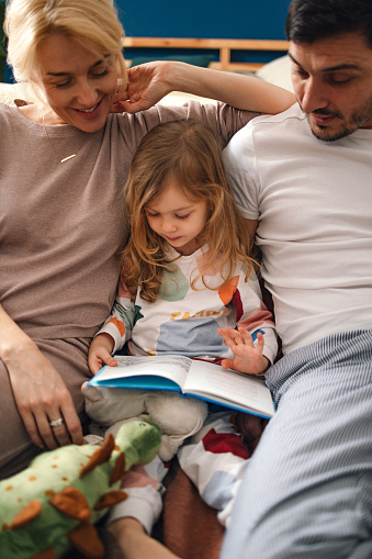 Happy little girl reading a fairy tale while sitting with her mother and father on the bedroom floor.