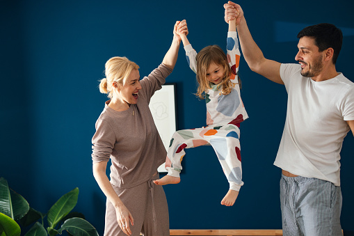 Smiling mother and father in sleepwear raising up and playing with their daughter in the bedroom.