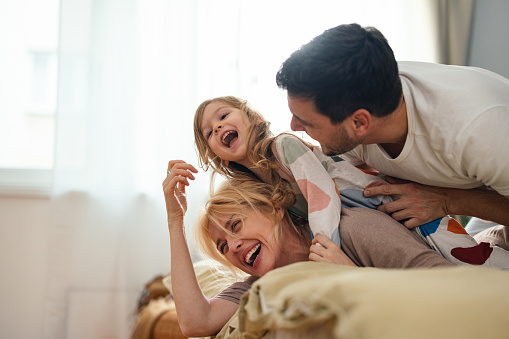 Smiling father and mother laughing and playing with their daughter while lying on a bed.