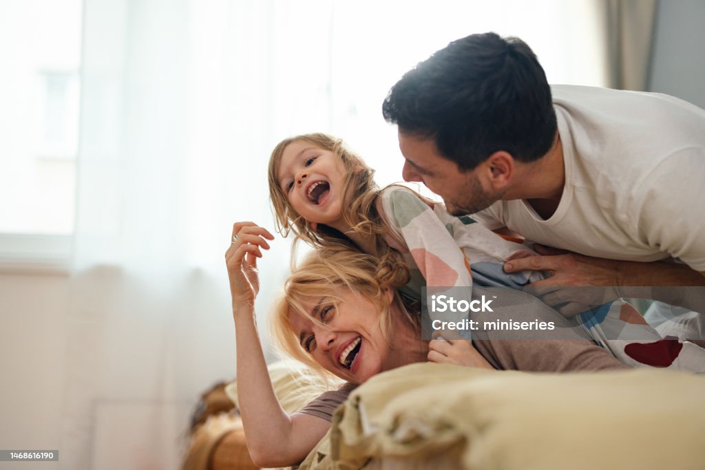 Happy Family In Sleepwear Having Fun Together In The Bedroom Smiling father and mother laughing and playing with their daughter while lying on a bed. Family Stock Photo
