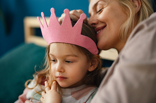 Smiling mother and her daughter with a princess crown lying on the bed and having fun together.