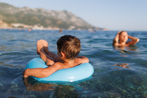 The baby sits in a blue inflatable small circle and looks for his mother, swimming in the blue warm summer Adriatic Sea in the evening sunlight