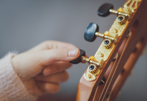 A little caucasian girl holds a guitar and twists the peg with her fingers while tuning the strings, close-up side view. Music education concept.