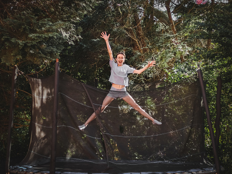 A beautiful slender caucasian teenage girl in shorts and a blouse jumps on a trampoline, frozen in a funny position of a star in the garden of a house among trees, close-up side view. Concept at home, home sports, happy childhood, children entertainment.