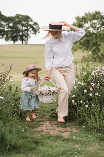 Mother and little daughter in field of daisie Cute little blonde girl in a cotton dress and straw hat walks in a field of daisies collects them in the basket with mum.