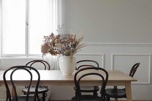Elegant dining room, scandinavian interior. Vase with dry flowers on wooden table. Vintage chairs. White wall moulding background, stucco decor and window. Empty copy space, elegant flat, home.