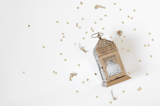 Festive Ramadan holiday composition, web banner. Golden ornamental Moroccan lantern and star shaped confetti isolated on white table background. Iftar party, Eid ul Fitr or Eid al Adha flatlay, top.