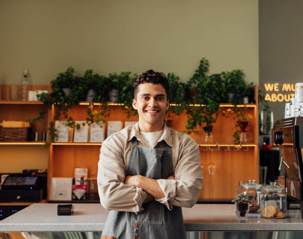 Coffee shop owner in an apron standing with crossed arms and smiling. Young handsome man leaning counter looking at a camera in a coffee shop. stock photo