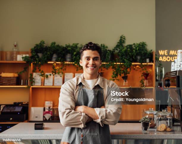 Coffee Shop Owner In An Apron Standing With Crossed Arms And Smiling Young Handsome Man Leaning Counter Looking At A Camera In A Coffee Shop Stock Photo - Download Image Now