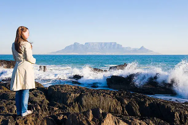 A girl standing on some rocks on the sea shore while waves are breaking gently over the rocks. Table Mountain in Cape Town, South Africa, is in the background.