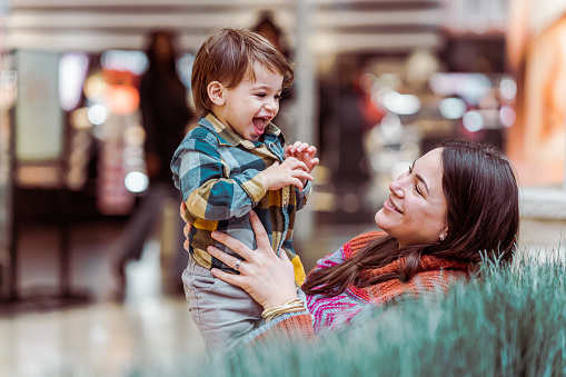 A beautiful woman of Puerto Rican descent sits on a bench inside a shopping mall and playfully interacts with her one year old toddler son who is laughing.