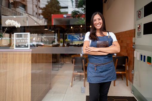 Portrait of a smiling female cafe owner standing in apron with arms crossed at open front door and looking at camera.