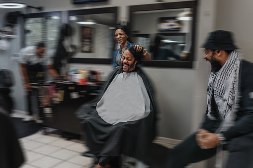 A black man who is at a barber shop having his hair braided laughs while talking with his female hair stylist and other customers.