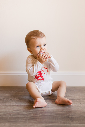 A Cute 13-Month-Old Baby Boy Barefoot, Sitting on the Floor & Playing with His Teether Toy, Wearing a Cloth Diaper Under a White Long-Sleeve Bodysuit That Says the Words 