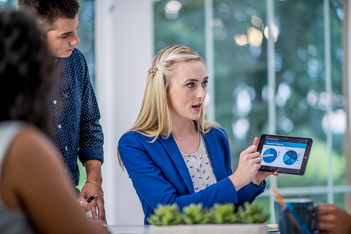 A young business woman holds out a tablet with pie charts ad recent stats on it, to her colleagues, as she presents the quarterly report.  She is dressed professionally and appears confident as she relays the information to the other partners.