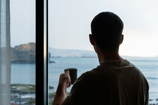 Asian man looking out a window and drinking coffee back view, concept of vacation or travel.