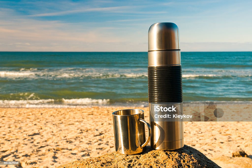 Thermos and metal cup Shiny metal thermos and a cup staying on a stone in front of the sea Beach Stock Photo