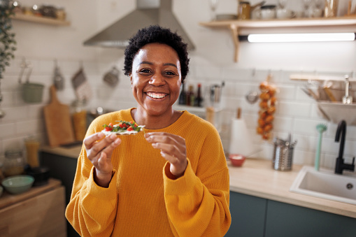 Portrait of young smiling beautiful woman in a domestic kitchen, holding cracker with vegetables in her hand