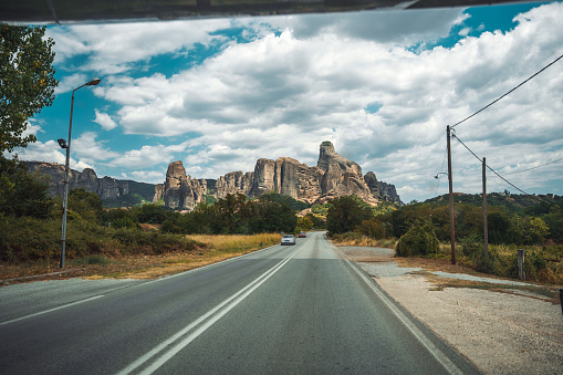 A view from inside the car of a road, leading to magnificent cliffs of Meteora with clouds in the background. Landscape image, another car approaching.