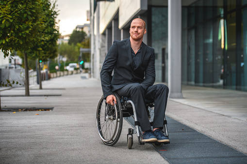 Full length front view of an attractive Caucasian male in a wheelchair. He is outdoors in a business center. He is looking away and smiling.