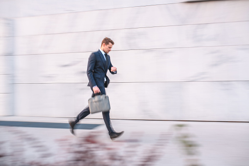 Full length side view of a mid adult Caucasian businessman hurrying to a meeting in a financial district. He is moving fast, carrying a briefcase and checking the time.