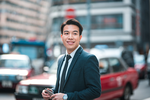 Smart young Japanese businessman commuting in a busy city. He is well-dressed, wearing a black suit and a tie. He is smiling and looking away.