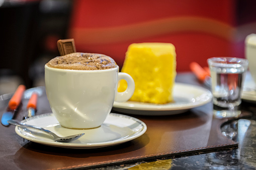 Cup of hot chocolate on a coffee shop table, with a slice of Brazilian couscous in the blurred background.
