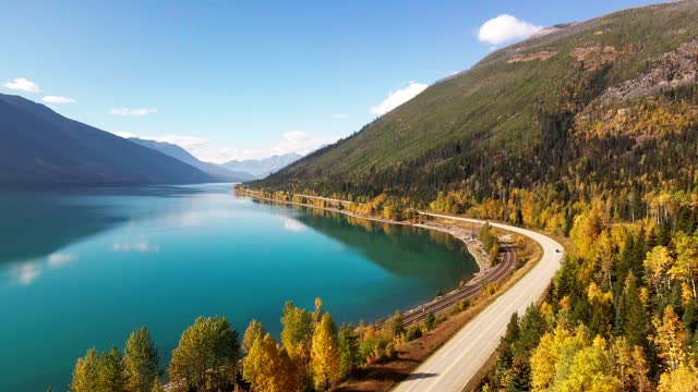 pan right aerial wide shot of Yellowhead Highway along moose lake in Mount Robson Provincial Park in autumn with yellow trees and mountains in the background and cars driving on the highway
