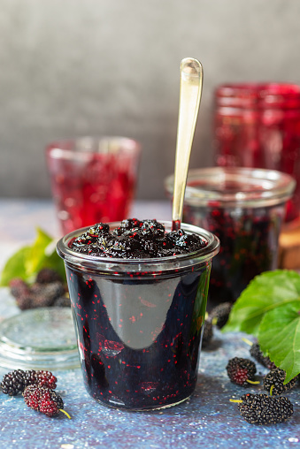 homemade mulberry jam in a glass jar with fresh mulberry with green leaves on rustic table background.