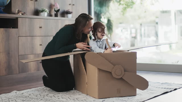 Mother little child son having fun playing with the plane from cardboard at home